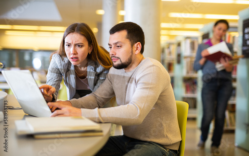 Positive man and woman spending time together in library, talking, work on laptop