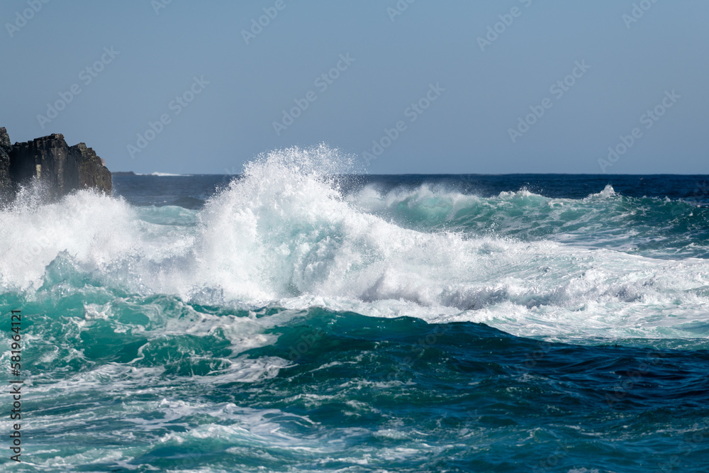 An angry turquoise green color massive rip curl of a wave as it barrels rolls along the ocean. The white mist and foam from the wave are foamy and fluffy. The ocean in the background is deep blue. 