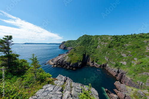 A rugged ocean coastline with high rocky sea stacks, cliffs, vibrant green grass, blue sky, and clouds. The smooth water is a blue color. There's a hiking trail along the edge of the cliff and coast. 