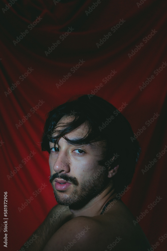 Fototapeta premium Artistic photo of a young latin man looking at camera with bare shoulders and a red cloth background. Vertical photo.