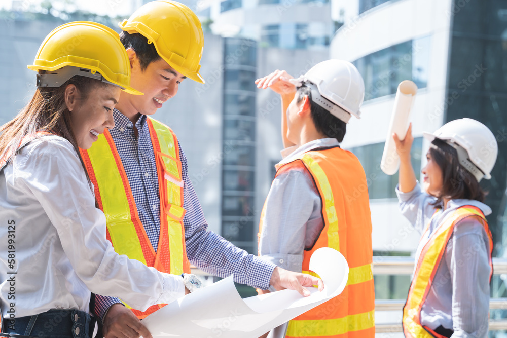 Group of Asian engineers in helmets looking at a blueprint to discuss and plan for build a modern building in city at construction site. Selective focus on engineering woman Group of Asian engineers in helmets looking at a blueprint to discuss and plan for build a modern building in city at construction site. Selective focus on engineering woman