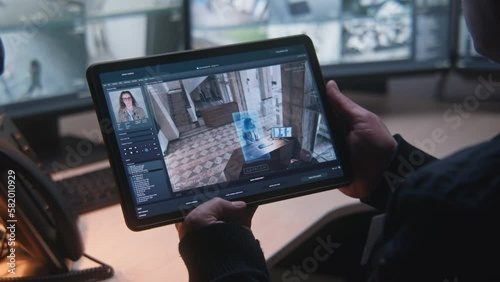 Male security officer controls CCTV cameras in office, uses digital tablet and computers with surveillance camera footage on screens. High tech security with face scanning. Tracking and social safety.