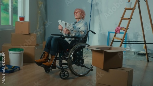 An elderly disabled woman in a wheelchair looks through the palette with colors. A granny plans repairs and chooses a paint color. Room with window, ladder, cardboard boxes, wallpaper, paint.