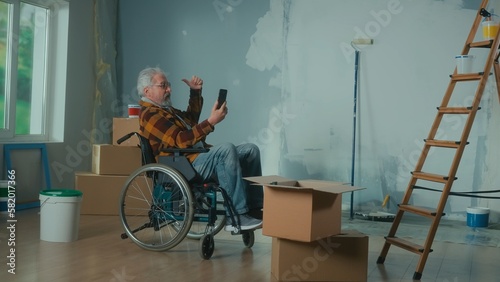 An elderly disabled man in a wheelchair talks on a video call using a mobile phone. A pensioner male points to the window. A room with window, wallpaper rolls, cardboard boxes and a bucket of paint.