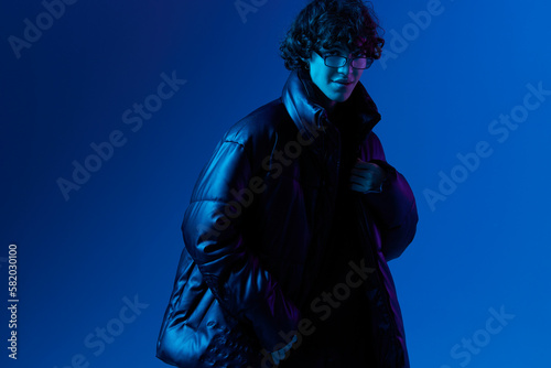 Man in winter down jacket model posing with glasses, hipster lifestyle, portrait blue background, mixed neon light, fashion style and trends guys teenager, copy space