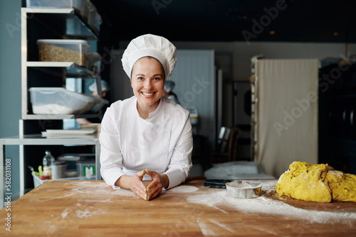 Portrait of positive baker in uniform leaned on wooden table with dough