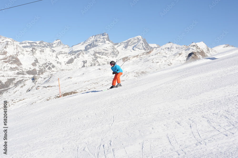 Skier in blue jacket, black helmet and orange pants on the piste slope in winter with snow mountains in Alps, Europe