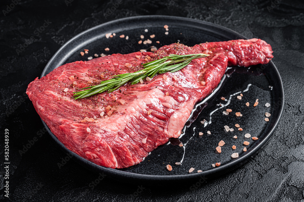 Seasoned raw tri-tip beef meat steak on plate. Black background. Top view