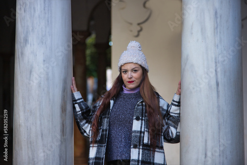 Young woman, beautiful, straight brown hair, leather pants, sweater, coat and wool hat, leaning on marble columns, looking at the camera, serious and worried. Concept fashion, autumn, winter, cold.