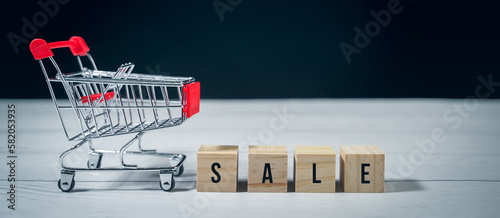 Sale word on wood block and empty shopping trolley cart on white wooden background. Concept retail e-commerce online store, promotion discount, black Friday, advertising for business consumer product.