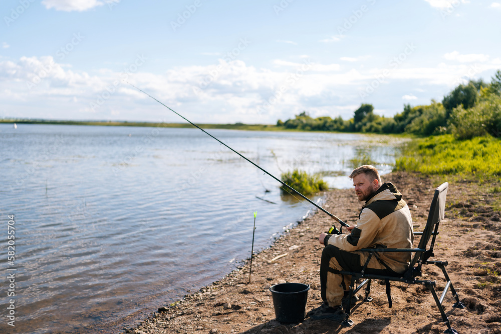Side view of bearded fisherman in raincoat sitting on river bank on travel chair with fishing rod waiting for catch on summer sunny day sandbank. Concept of lifestyle, leisure activity on nature Side view of bearded fisherman in raincoat sitting on river bank on travel chair with fishing rod waiting for catch on summer sunny day sandbank. Concept of lifestyle, leisure activity on nature