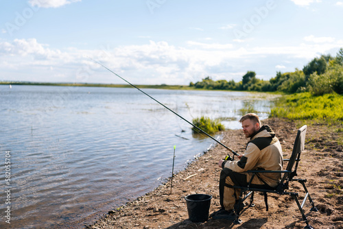 Side view of bearded fisherman in raincoat sitting on river bank on travel chair with fishing rod waiting for catch on summer sunny day sandbank. Concept of lifestyle, leisure activity on nature