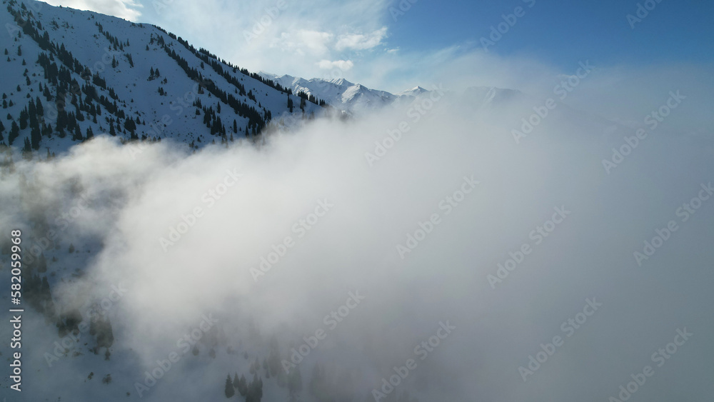 White cumulus clouds in snowy mountains in winter. The rays of the sun fall on part of the clouds, a shadow comes from the peak. Blue clear sky. Christmas trees grow on the hills. Tourists are walking
