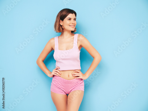 Portrait of young beautiful smiling female in trendy summer shorts clothes. Carefree woman posing near blue wall in studio. Sexy positive model having fun indoors. Cheerful and happy © halayalex