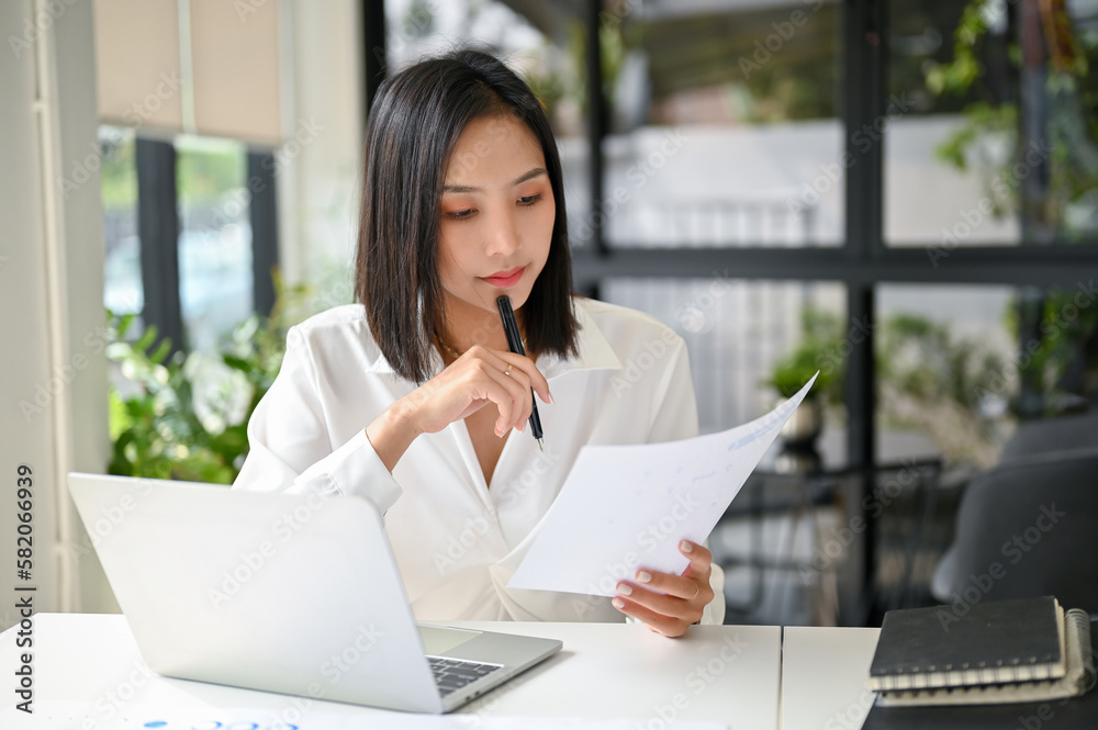 Concentrated Asian businesswoman reviewing or examining business reports at her desk.