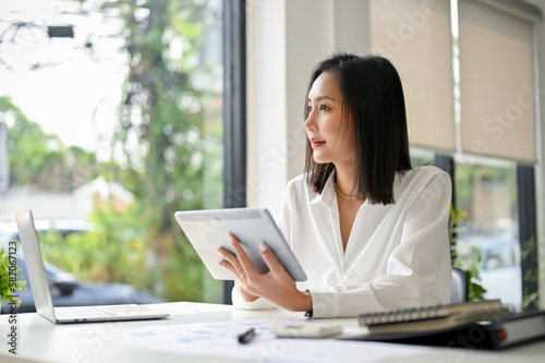 Gorgeous Asian businesswoman using her tablet, looking out the window, pondering and daydreaming