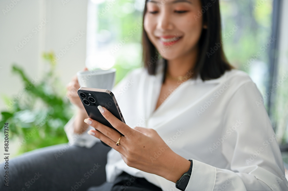 Smiling Asian businesswoman using her phone and having coffee in her office. Smiling Asian businesswoman using her phone and having coffee in her office.