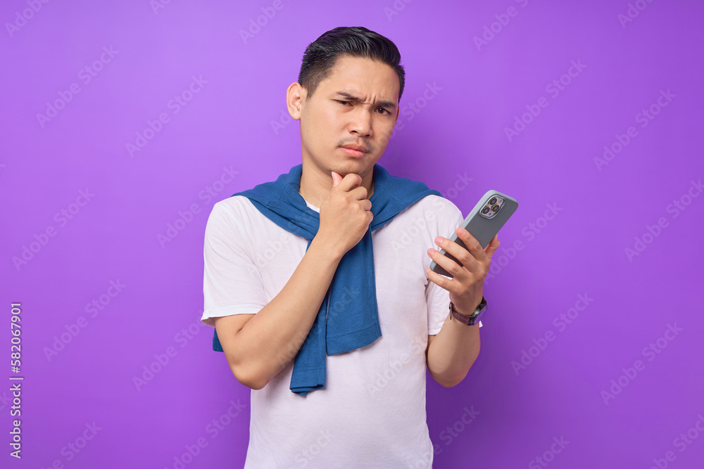 Pensive young Asian man in casual clothes holding smartphone and looking at camera, thinking about offer isolated on purple background Pensive young Asian man in casual clothes holding smartphone and looking at camera, thinking about offer isolated on purple background