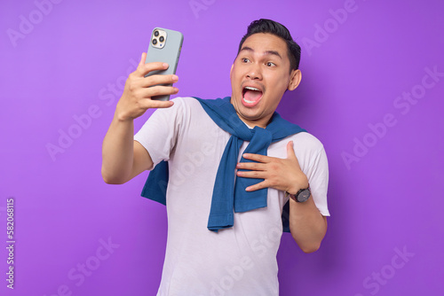 Surprised young Asian man using smartphone, reading special offer isolated over purple background © Bangun Stock Photo