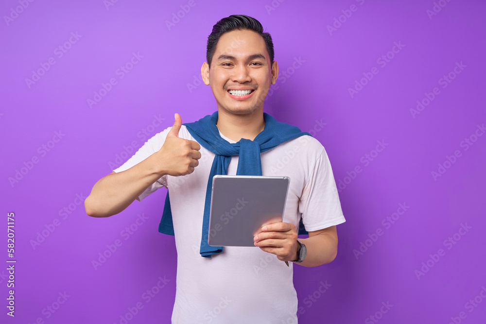 Smiling young Asian man wearing t-shirt casual clothes holding digital tablet and showing thumb up gesture isolated over purple background