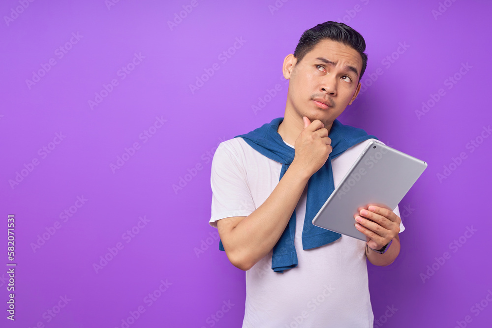 Portrait of pensive young Asian man in white t-shirt touching chin and holding digital tablet isolated on purple background Portrait of pensive young Asian man in white t-shirt touching chin and holding digital tablet isolated on purple background