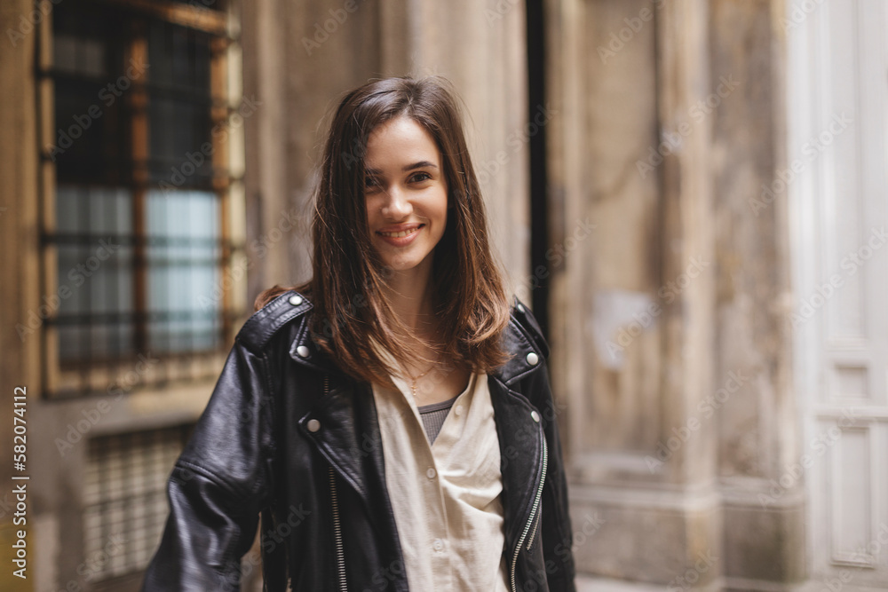 Trendy girl with brunette hairstyle posing outside. Woman in leather jacket and shirt walking on the street outdoors. Tourist happy woman posing in the city. Optimistic lady turn around.