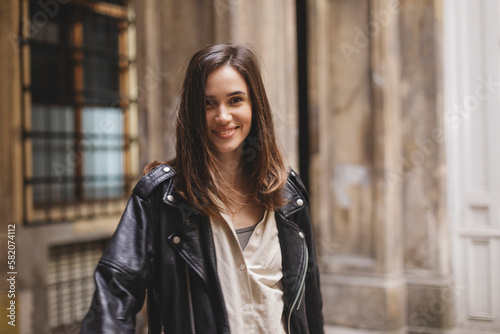 Trendy girl with brunette hairstyle posing outside. Woman in leather jacket and shirt walking on the street outdoors. Tourist happy woman posing in the city. Optimistic lady turn around.