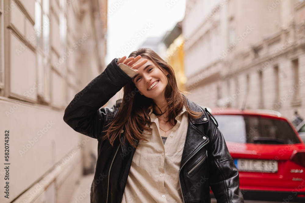 Fototapeta premium Trendy girl with brunette hairstyle posing outside. Woman in leather jacket and shirt walking on street outdoors. Tourist happy woman posing in the city. Optimistic lady walks outside, hand near face.
