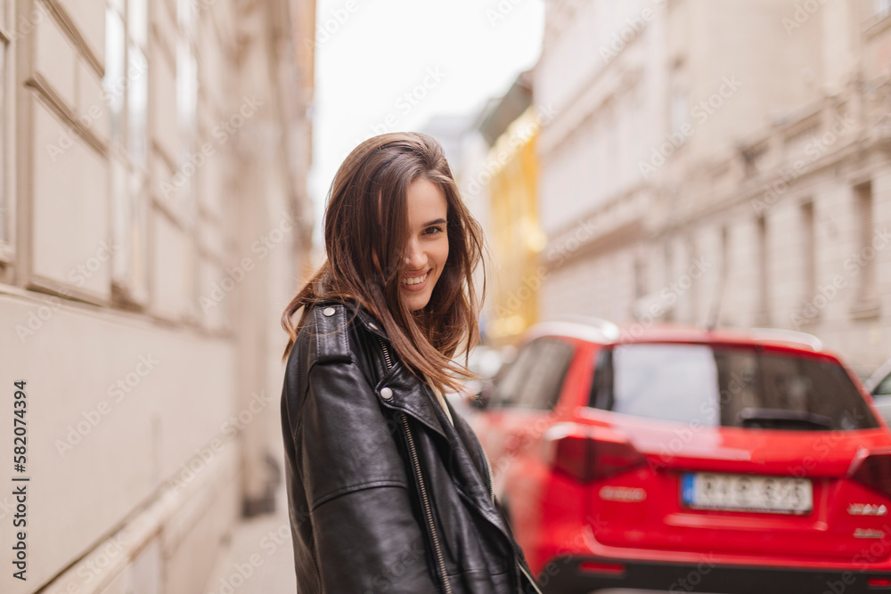 Trendy girl with brunette hairstyle posing outside. Woman in leather jacket and shirt walking on the street outdoors. Tourist happy woman posing in the city. Optimistic lady turn around.