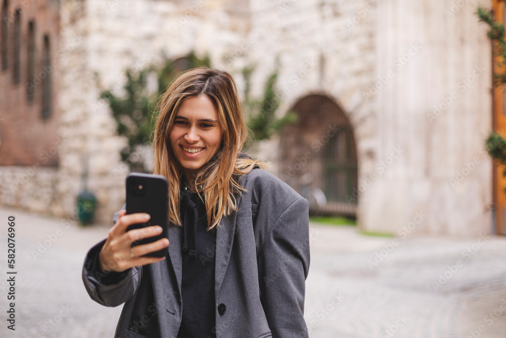 Happy young woman photographing herself using her mobile phone. Caucasian female talking selfie with her smart phone walking outdoor on the street. Girl posing in grey suit, t-shirt.