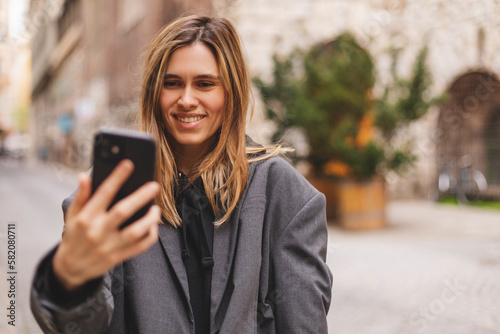 Happy young woman photographing herself using her mobile phone. Caucasian female talking selfie with her smart phone walking outdoor on the street. Girl posing in grey suit, t-shirt.