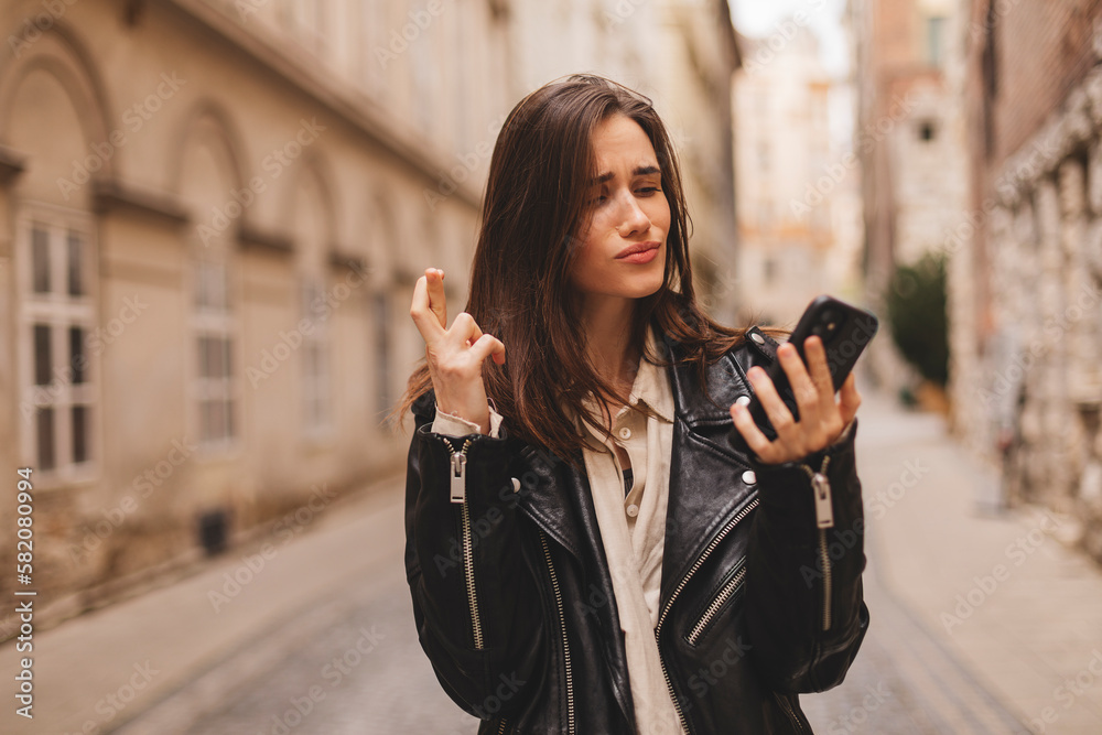 Fototapeta premium Portrait of wishful young woman in casual clothes with brunette hair, crossing her fingers, feeling nervous before important event, walking on the street. Body language.