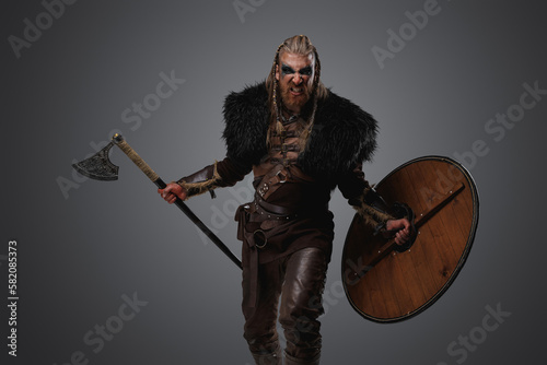 Studio shot of screaming nordic warrior with black fur and axe isolated on grey background.