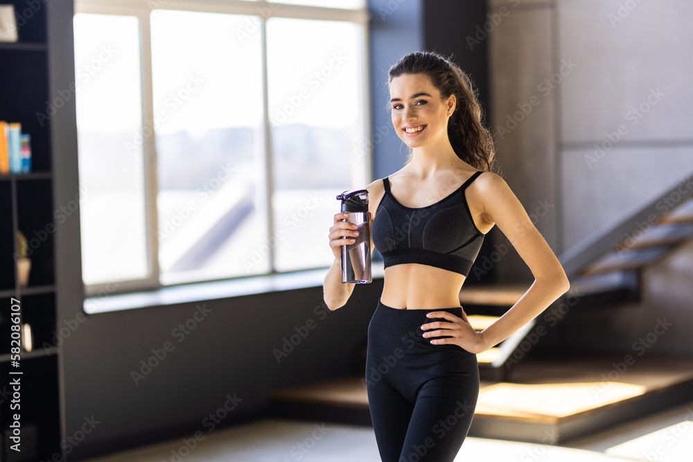 Portrait of sporty beautiful smiling lady in sportswear and white towel on neck holding shaker with healthy drink, whey protein, fresh water or coffee