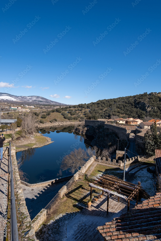 Paisaje del río de Lozoya rodeando el pueblo madrileño Buitrago de Lozoya con las montañas del fondo desenfocadas bajo un cielo azul soleado.
