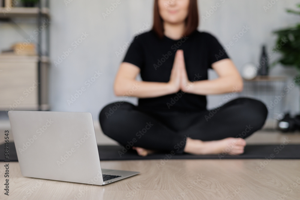 Naklejka premium sport and internet concept - young woman sitting on yoga mat and doing yoga with online tutorial on laptop