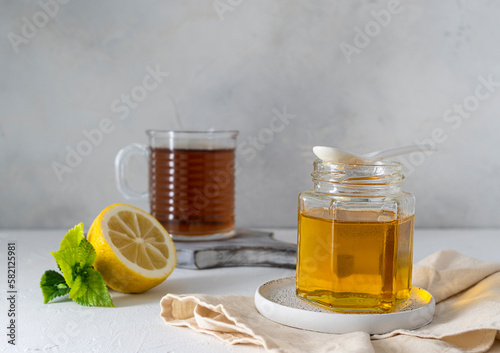 Floral honey in a transparent jar on a light table. A white ceramic spoon on a jar. Tea in a glass glass and half a lemon in the background. Light background