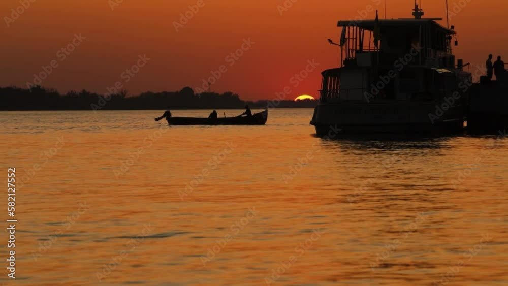 The disk of the sun goes over the horizon over the Danube River, the dark silhouettes of fishermen on a rowboat and a pleasure boat against the crimson sunset