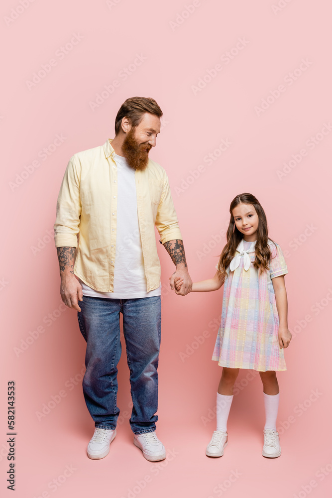 Full length of tattooed man holding hand of preteen daughter on pink background. Full length of tattooed man holding hand of preteen daughter on pink background.