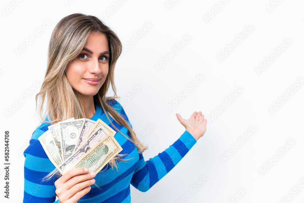 Young Uruguayan woman taking a lot of money isolated on white background extending hands to the side for inviting to come Young Uruguayan woman taking a lot of money isolated on white background extending hands to the side for inviting to come