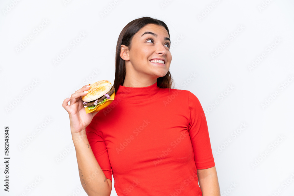 Young caucasian woman holding a burger isolated on white background looking up while smiling Young caucasian woman holding a burger isolated on white background looking up while smiling