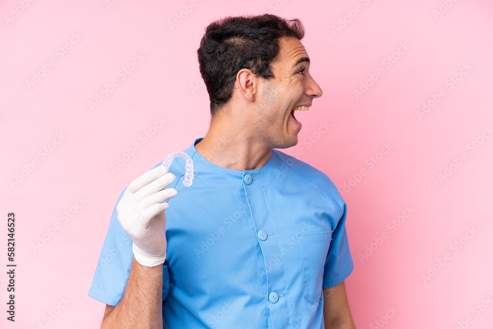 Dentist caucasian man holding invisible braces isolated on pink background laughing in lateral position Dentist caucasian man holding invisible braces isolated on pink background laughing in lateral position