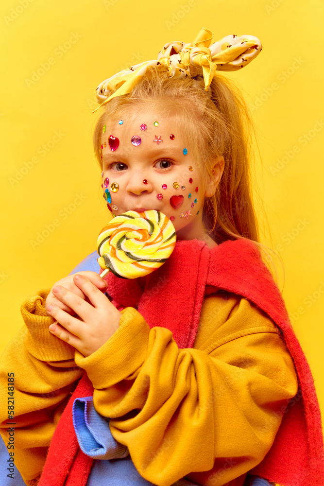 Photo of adorable little girl holding lollipop and eating sweet candy over yellow background. Concept of fashion, ad, children's products, child model, clothing and accessories