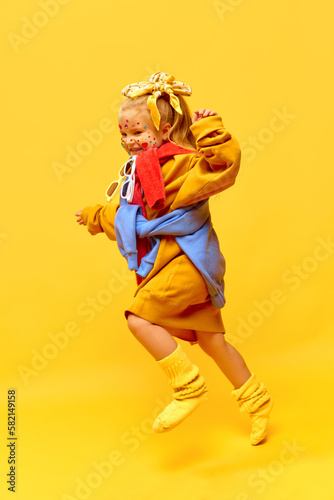 Full-length profile photo of running away little, happy girl in funny oversized clothes over yellow background. Happiness photo