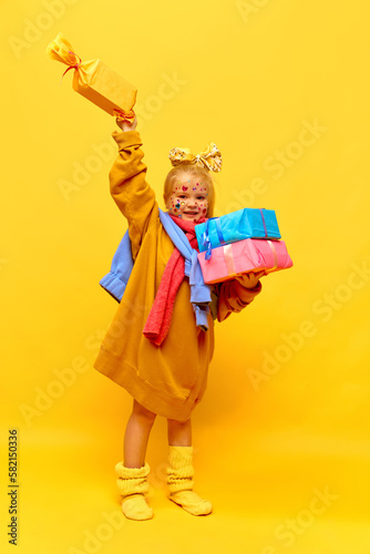 Full-length photo of little girl holding gifts, showing box with happy face over yellow background. Concept of celebration, ad, children's products, childhood, family photo