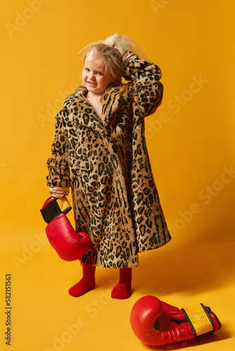 Little blond girl wearing boxing gloves and leopard furcoat standing and looking away over yellow background. Full-length photo