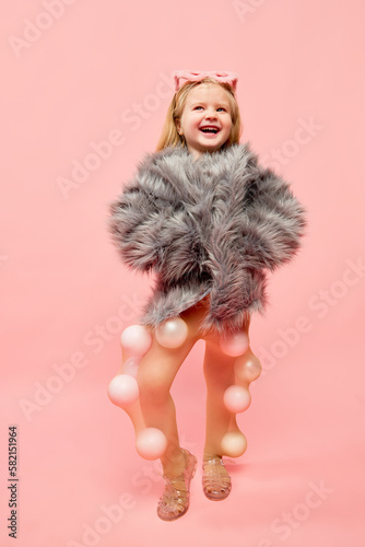 Photo of little happy girl wearing sunglasses and fur coat, keeping hands in pocket with happy face looking away over pink background