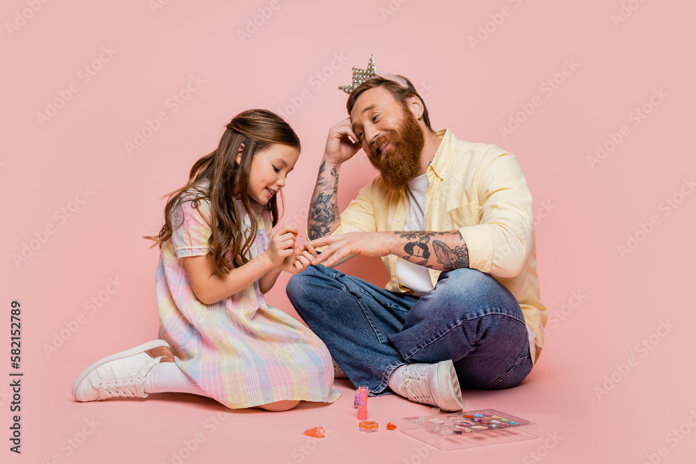 Cheerful girl applying nail polish on hand of tattooed dad with crown headband on pink background.