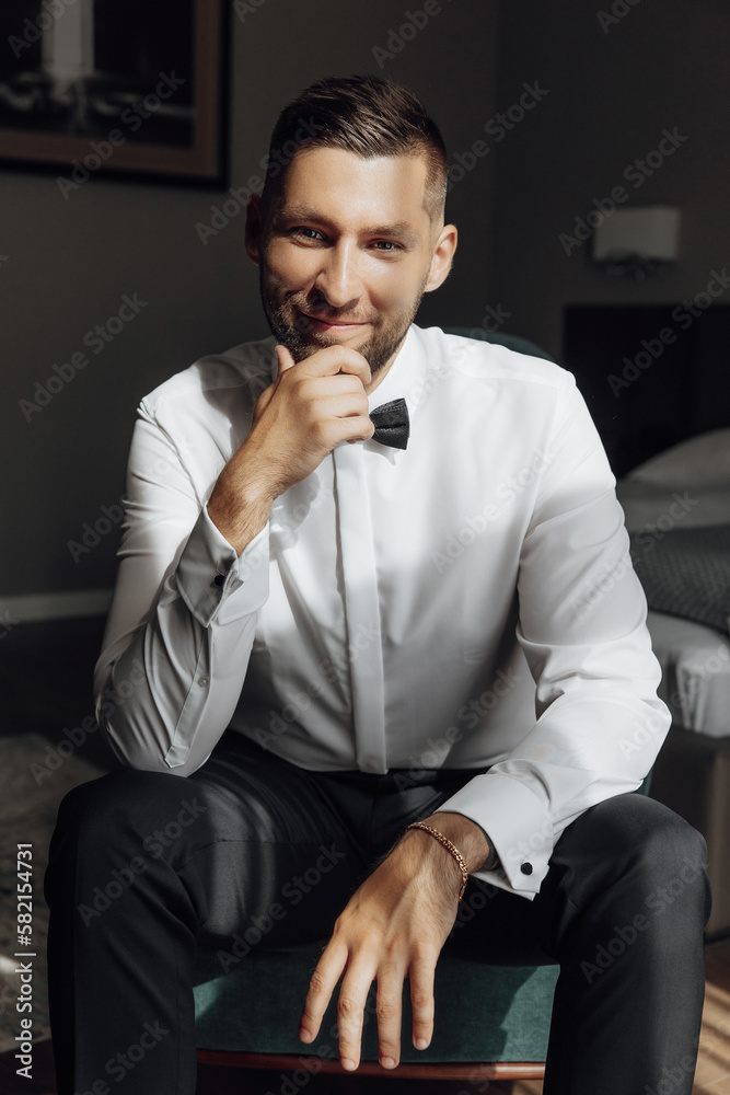 The stylish bridegroom poses looking at the camera, preparing for a wedding ceremony. Large portrait. Wonderful interior in the morning light. The groom's morning The stylish bridegroom poses looking at the camera, preparing for a wedding ceremony. Large portrait. Wonderful interior in the morning light. The groom's morning