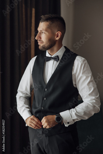 Morning of a stylish groom, man, in a black suit and tie, his preparations at the hotel, in a photo studio, fastening buttons and jacket, portraits